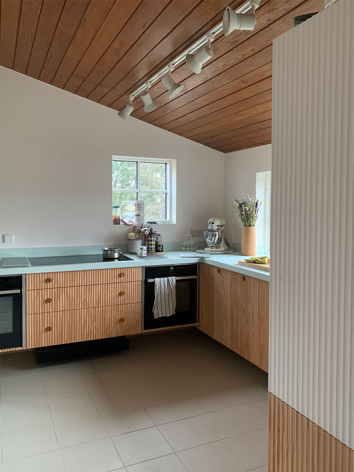 Furthering the seamless integration of the new kitchen design into the existing structure of the house, the Douglas Fir clad ceiling is mirrored in the choice to use the same timber for the fluted kitchen cabinets.
